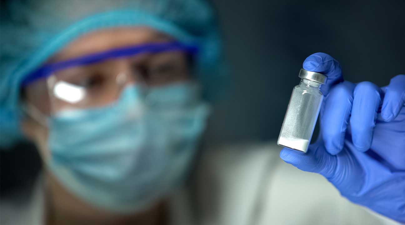 doctor wearing a mask and gloves holding a small vile of potent carfentanil