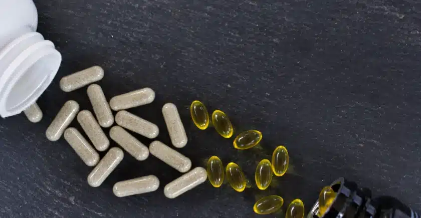 Top view of scattered white capsule pills and yellow softgel capsules on a black surface, with partially visible bottles