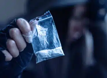 Man in black hoodie holding a dime bag of a white powder substance.